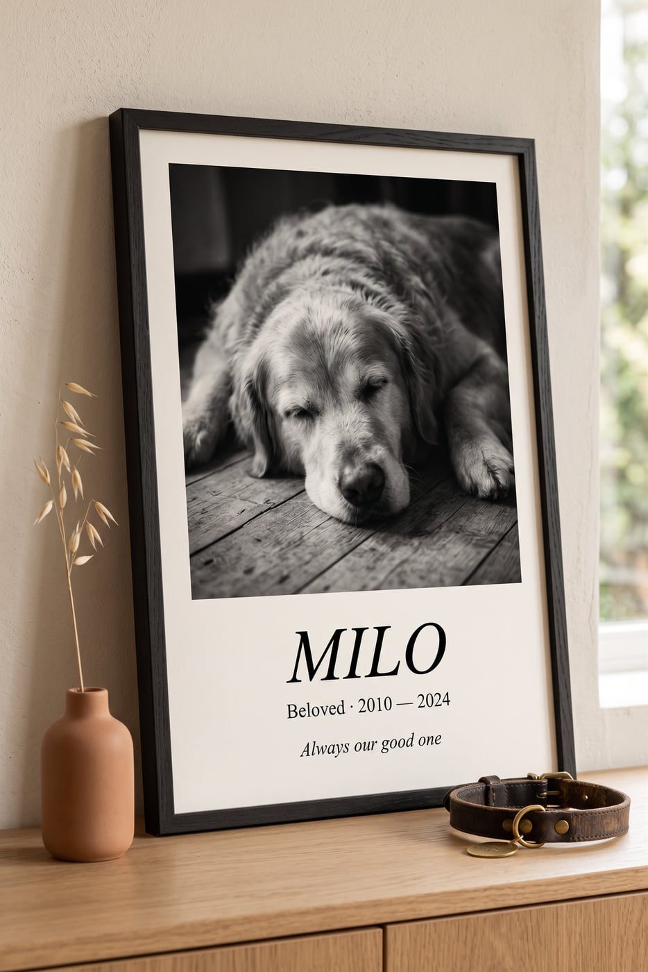 A framed black-oak memorial poster on an oak console table showing a black-and-white photo of a golden retriever, with the caption MILO · Beloved 2010–2024 · Always our good one